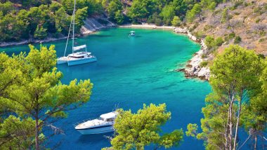 La tranquilla baia di Brac in Croazia con acque cristalline, circondata da lussureggiante vegetazione verde. Due barche a vela, una più grande con un catamarano bianco e una più piccola, sono ancorate nel mare calmo, invitando a una rilassante vacanza in barca a vela in Croazia. Il panorama promette un'avventura unica per le vacanze in barca a vela, ideale per chi desidera esplorare le incantevoli coste croate con una crociera in barca a vela.