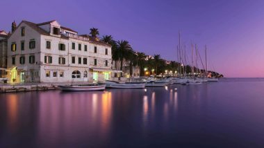 Citta di Hvar vista lungomare, Tramonto serale su piccolo porto con edifici storici in pietra chiara e palme, barche a vela ormeggiate in fila lungo la banchina riflettendosi sulle acque calme e violacee del mare.