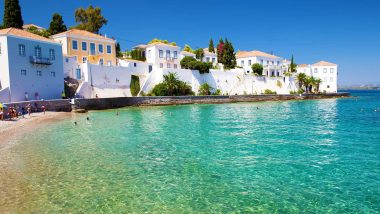 Piccola Baia dell'Isola di Spetses - In questa vivida fotografia si può ammirare una spiaggia incantevole delle Isole del Saronico, con acque cristalline che bagnano una sabbia fine. In lontananza, una serie di case tradizionali greche con le loro facciate dipinte di bianco e i dettagli in colori vivaci creano una pittoresca scena mediterranea.