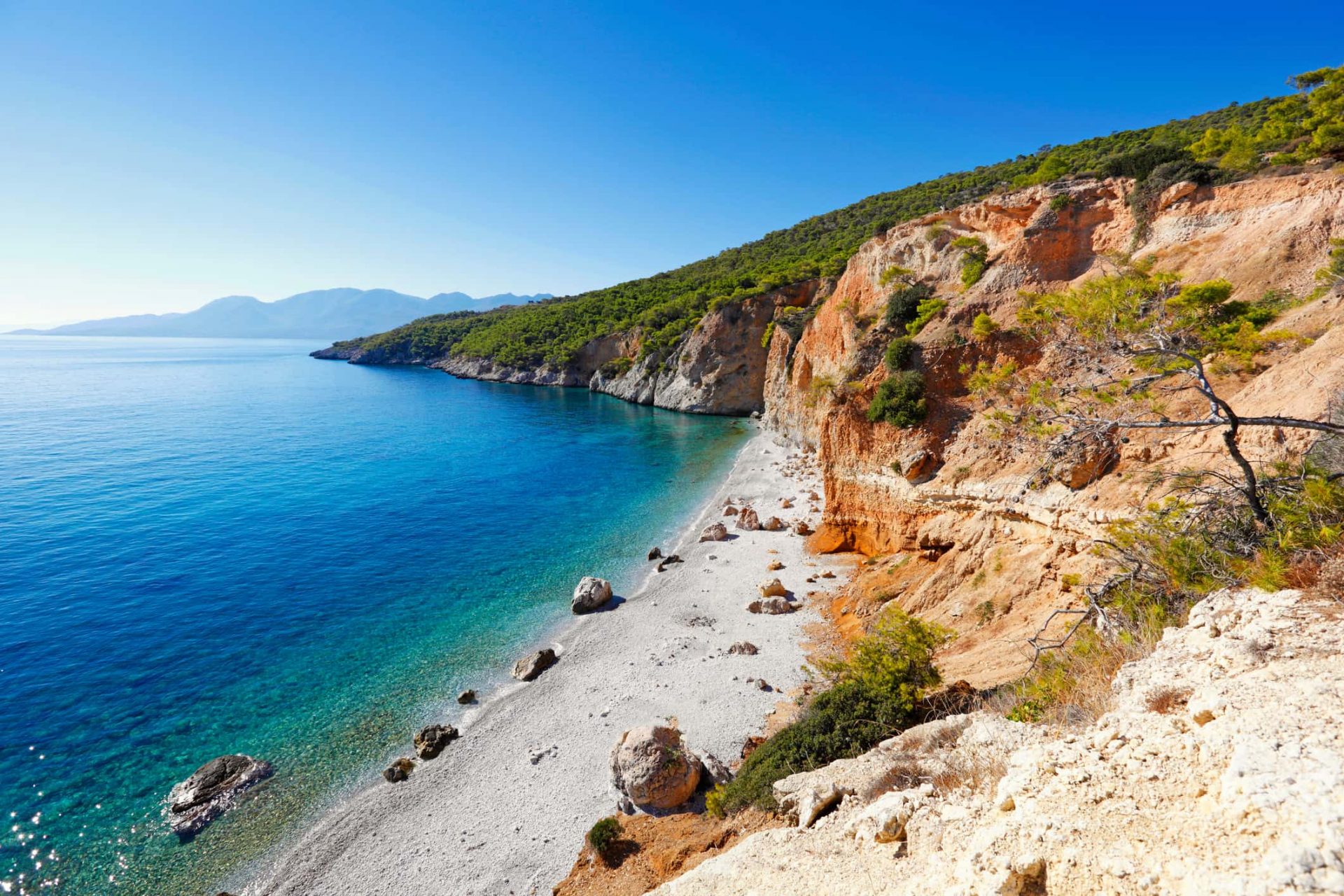 Spiaggia di Chialikiada Isola Angistri caratterizzata da un impressionante dirupo roccioso che scende a picco nel mare cristallino. La riva è un mix di sassolini bianchi e grandi rocce sparse, le cui tonalità calde contrastano con il blu intenso e puro dell'acqua.