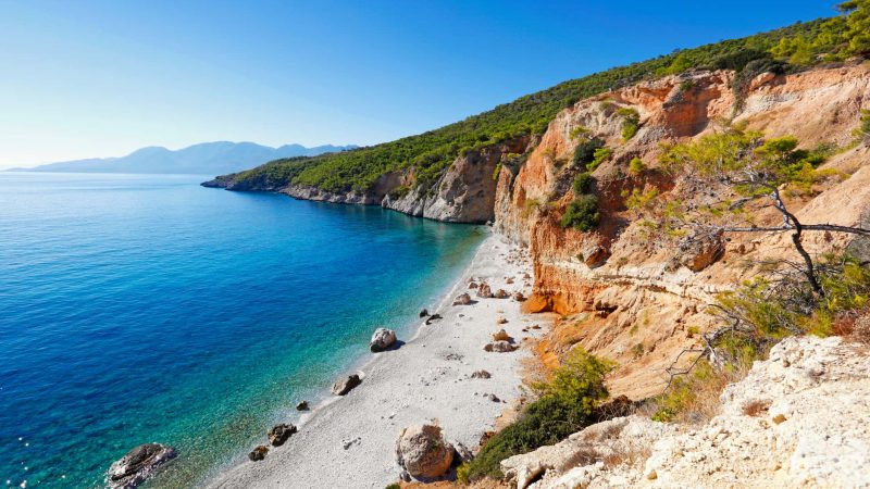 Spiaggia di Chialikiada Isola Angistri caratterizzata da un impressionante dirupo roccioso che scende a picco nel mare cristallino. La riva è un mix di sassolini bianchi e grandi rocce sparse, le cui tonalità calde contrastano con il blu intenso e puro dell'acqua.