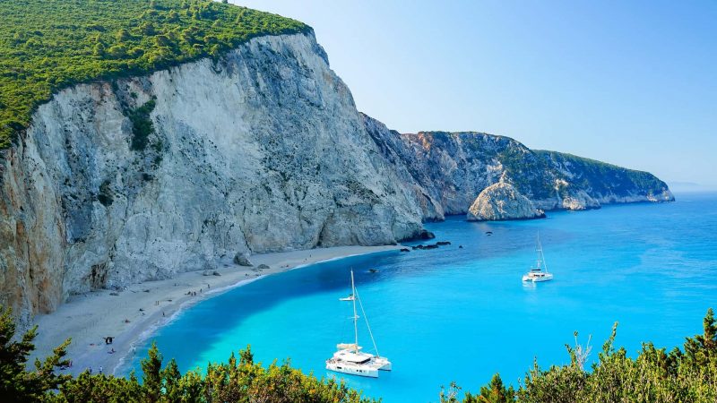 Catamarano bianco ancorato nelle acque turchesi presso la Spiaggia di Porto Katsiki, Lefkada, con imponenti scogliere calcaree e vegetazione lussureggiante. Vacanze in Barca a Vela alle Isole Ionie