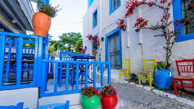 Un Vicolo sull'Isola di Rodi: angolo caratteristico di un caffè greco con sedie blu e gialle e fiori rossi che adornano le pareti bianche e blu.